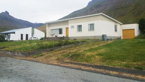 A modern two-story building with a flat roof, large windows, and a garage. It is situated in a grassy area with mountains in the background.