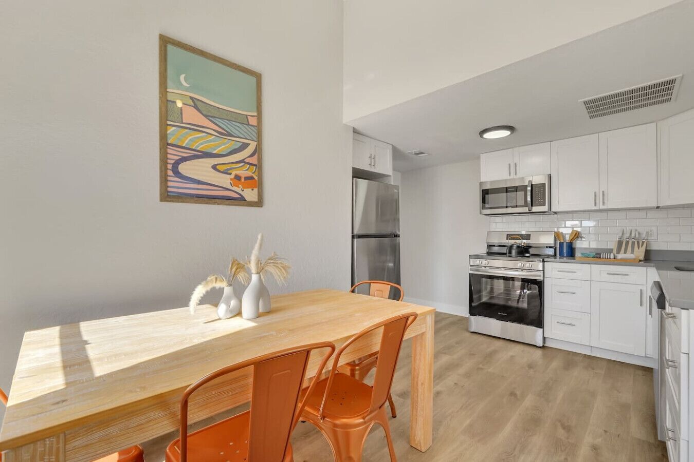 A modern kitchen with white cabinets, stainless steel appliances, and a wooden dining table with orange chairs.