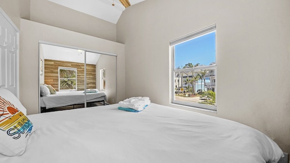Une chambre d’hôtel avec un lit, une grande fenêtre, un miroir et une vue sur un immeuble bordé de palmiers.