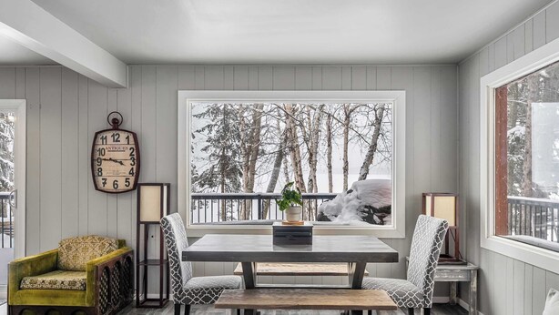A cozy room with a wooden table and chairs, a vintage clock, and a view of trees through a large window. - Summer Lake Cabin + Boat Slip + Fire Pit + Beach (Lac La Nonne)
