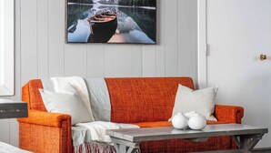 A room with a wooden panel wall, a framed picture of a mountain lake, an orange sofa with white and orange cushions, and a small table with white vases. - Summer Lake Cabin + Boat Slip + Fire Pit + Beach (Lac La Nonne)