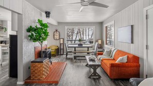 A modern living room with a dining area, a ceiling fan, a large window with a view of trees, and a fireplace. - Summer Lake Cabin + Boat Slip + Fire Pit + Beach (Lac La Nonne)