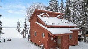 A wooden cabin with a snow-covered roof, surrounded by trees and a snowy landscape. - Summer Lake Cabin + Boat Slip + Fire Pit + Beach (Lac La Nonne)