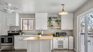 A modern kitchen with white cabinetry, a central island, a ceiling fan, and a view through a large window. - Summer Lake Cabin + Boat Slip + Fire Pit + Beach (Lac La Nonne)
