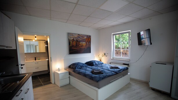 A modern bedroom with a bed, a small bedside table, a window, a TV, and a kitchen area visible through an open door. - Deluxe Double Studio with kitchen line, bathroom, Shower, Toilet & king bed (Rudolstadt)