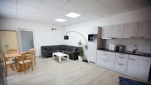A modern living room with a sofa, dining table, and chairs, a kitchen with cabinets and appliances, and a ceiling with recessed lighting. - Deluxe Double Studio with kitchen line, bathroom, Shower, Toilet & king bed (Rudolstadt)