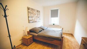 Ein Schlafzimmer mit Holzboden, einem Bett mit grauer Decke und Kissen, einem Nachttisch mit Lampe, einer Stehlampe und einem Fenster mit Blick nach draußen.