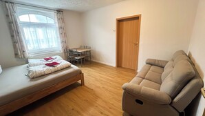 A small, well-lit room with a bed, a sofa, a dining table with chairs, and a door.