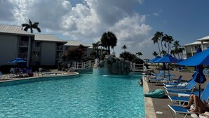 A pool area with lounge chairs and umbrellas, a rock feature, and a building with balconies.