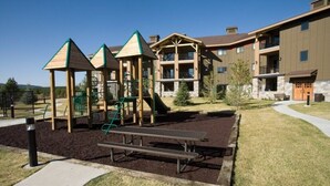 A playground with climbing structures and a picnic table, surrounded by a building with balconies and a stone facade. - WorldMark Yellowstone - 1 Bedroom (West Yellowstone)