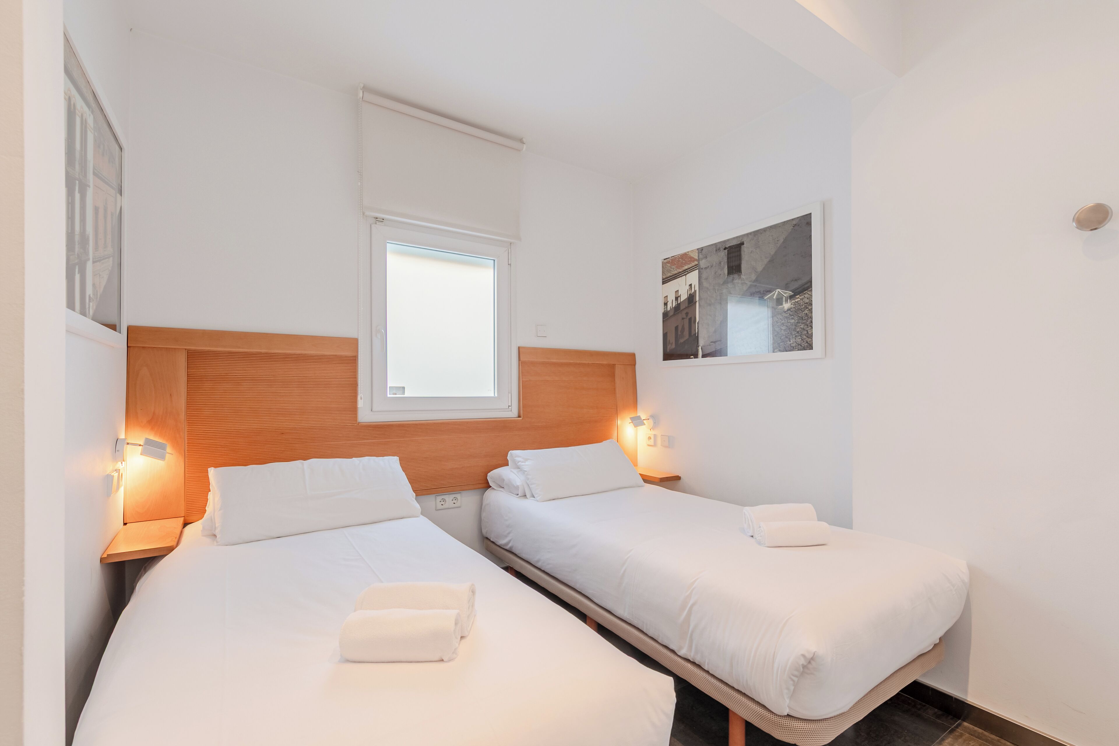 A hotel room with two single beds, a wooden headboard, bedside lamps, a window, and a framed picture on the wall.