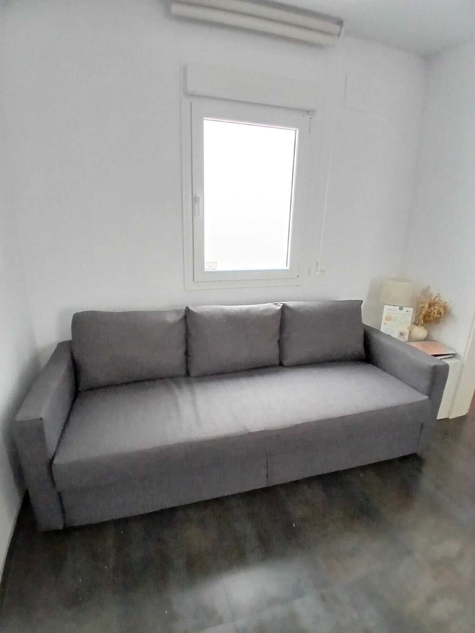 Um sofá cinzento numa sala com janela, uma mesinha de centro e uma luminária, e uma parede branca.