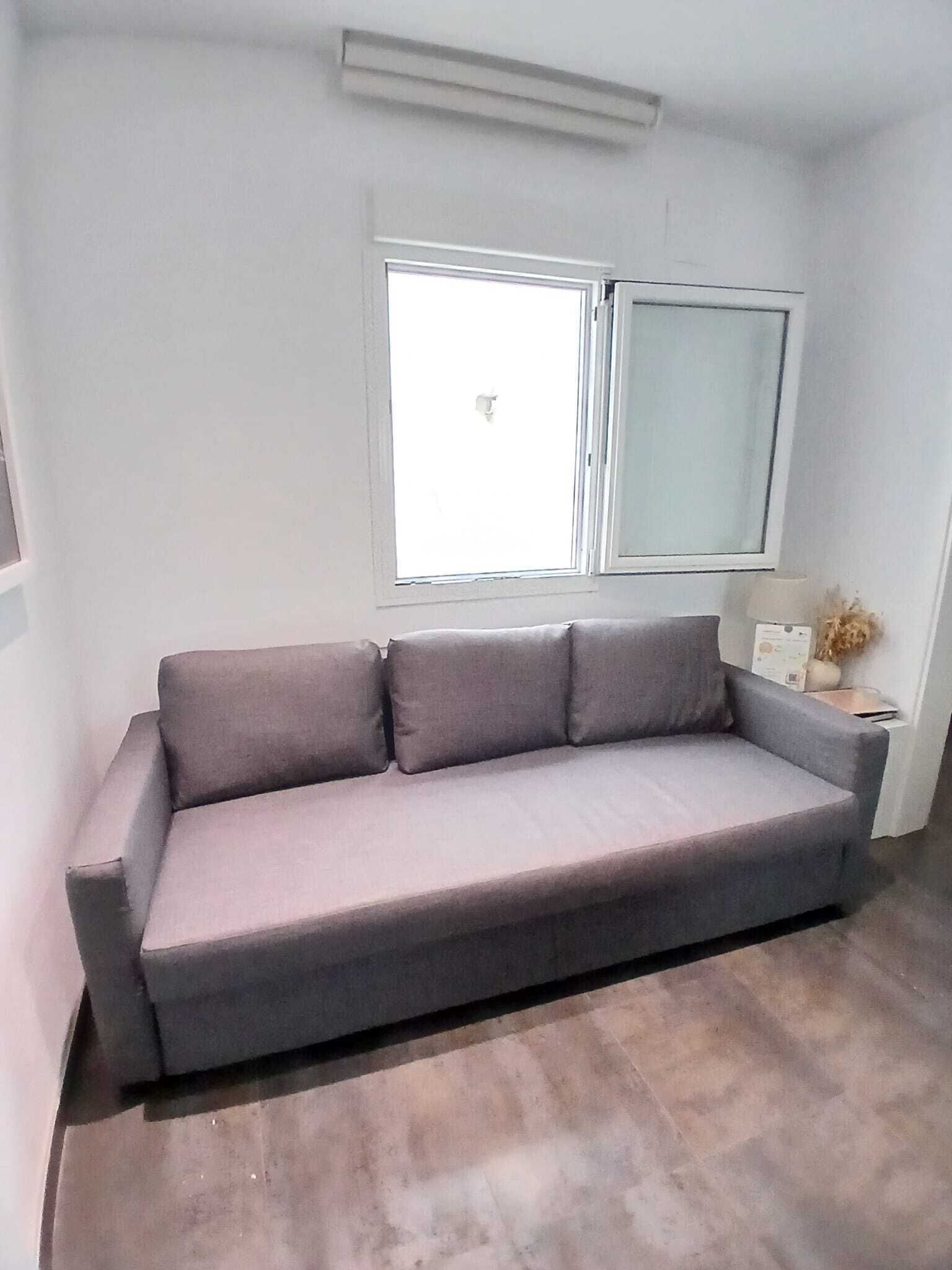 Un canapé gris dans une pièce avec une fenêtre, une lampe et une petite table sur laquelle se trouve un vase.