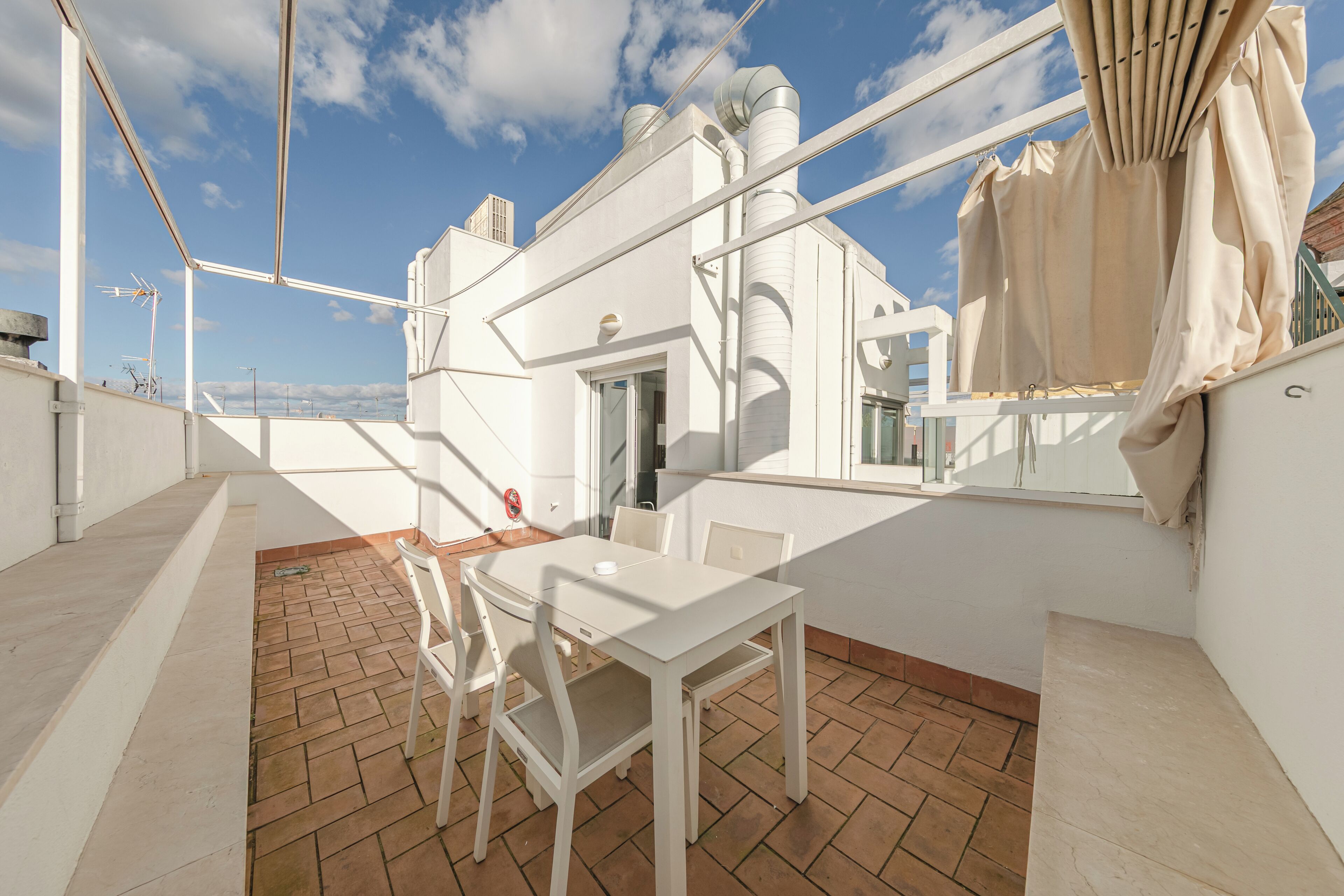 Eine Dachterrasse mit weißem Tisch und Stühlen, Blick auf die Stadt und ein Gebäude mit weißen Wänden und einem Schornstein.