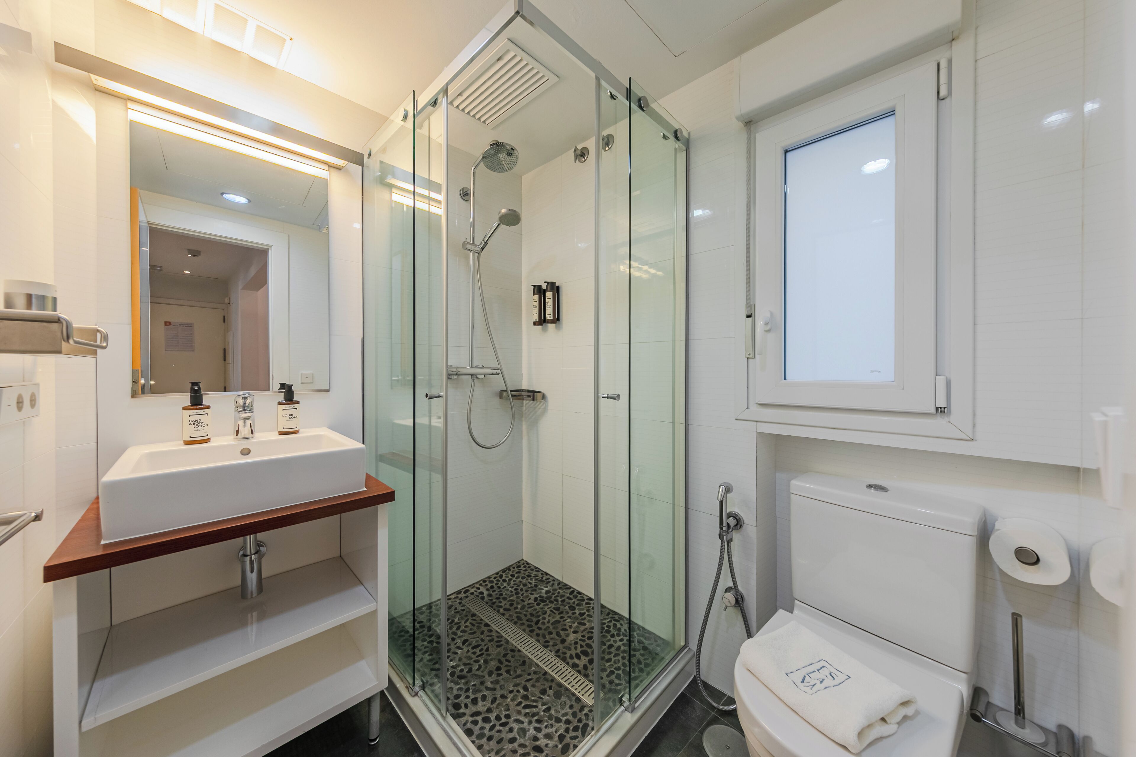 Een moderne badkamer met een douchecabine van glas, een wit wastafeltje met twee flesjes lotion en een toilet met een handdoek.