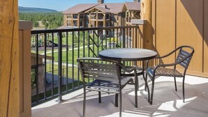 A balcony with a table and chairs, overlooking a building with multiple windows and a view of a green landscape. - Yellowstone | Adventure Base Stay | Pool + Nature (West Yellowstone)