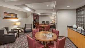 A hotel room with a dining area featuring a round wooden table and pink chairs, a sofa, a small side table with a lamp, and a view of the adjacent room. - Yellowstone | Adventure Base Stay | Pool + Nature (West Yellowstone)