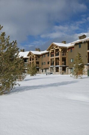 A multi-story lodge with a snow-covered roof and stone foundation, surrounded by snow and pine trees.