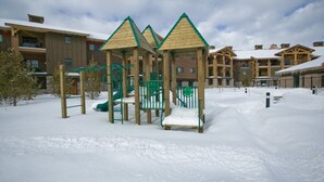 A snow-covered playground with climbing structures and slides, surrounded by multi-story buildings with a rustic architectural style. - Yellowstone | Adventure Base Stay | Pool + Nature (West Yellowstone)