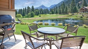 Un patio al aire libre con una parrilla, una mesa redonda y cuatro sillas. El patio da a un estanque con una fuente, rodeado de césped cuidado y árboles.