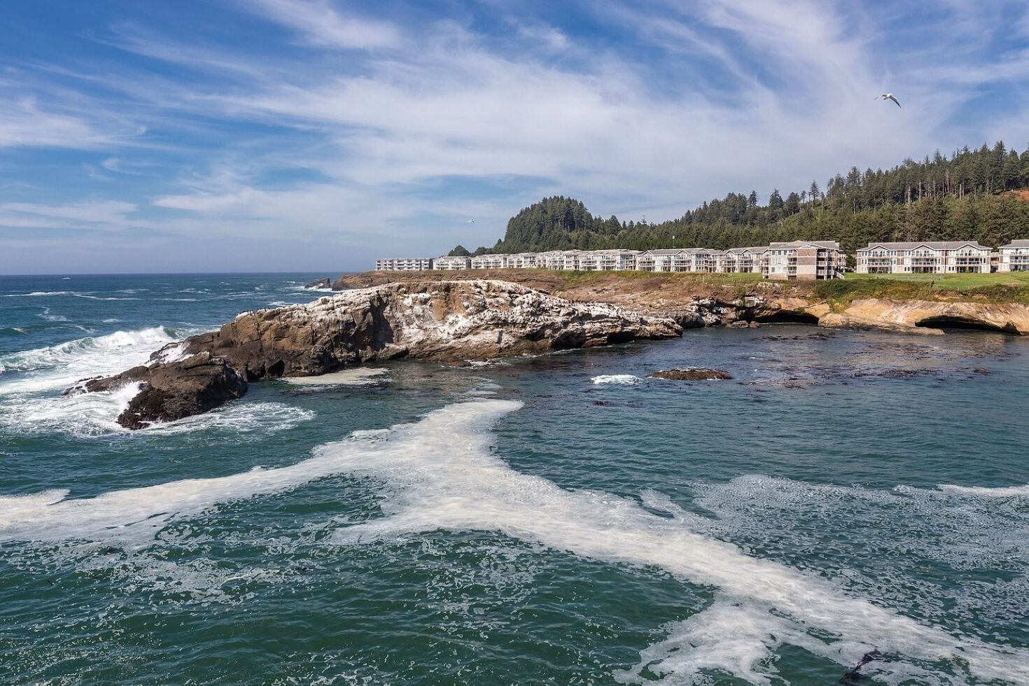 A coastal hotel with a rocky shoreline, waves crashing against the rocks, and a forested hillside in the background.