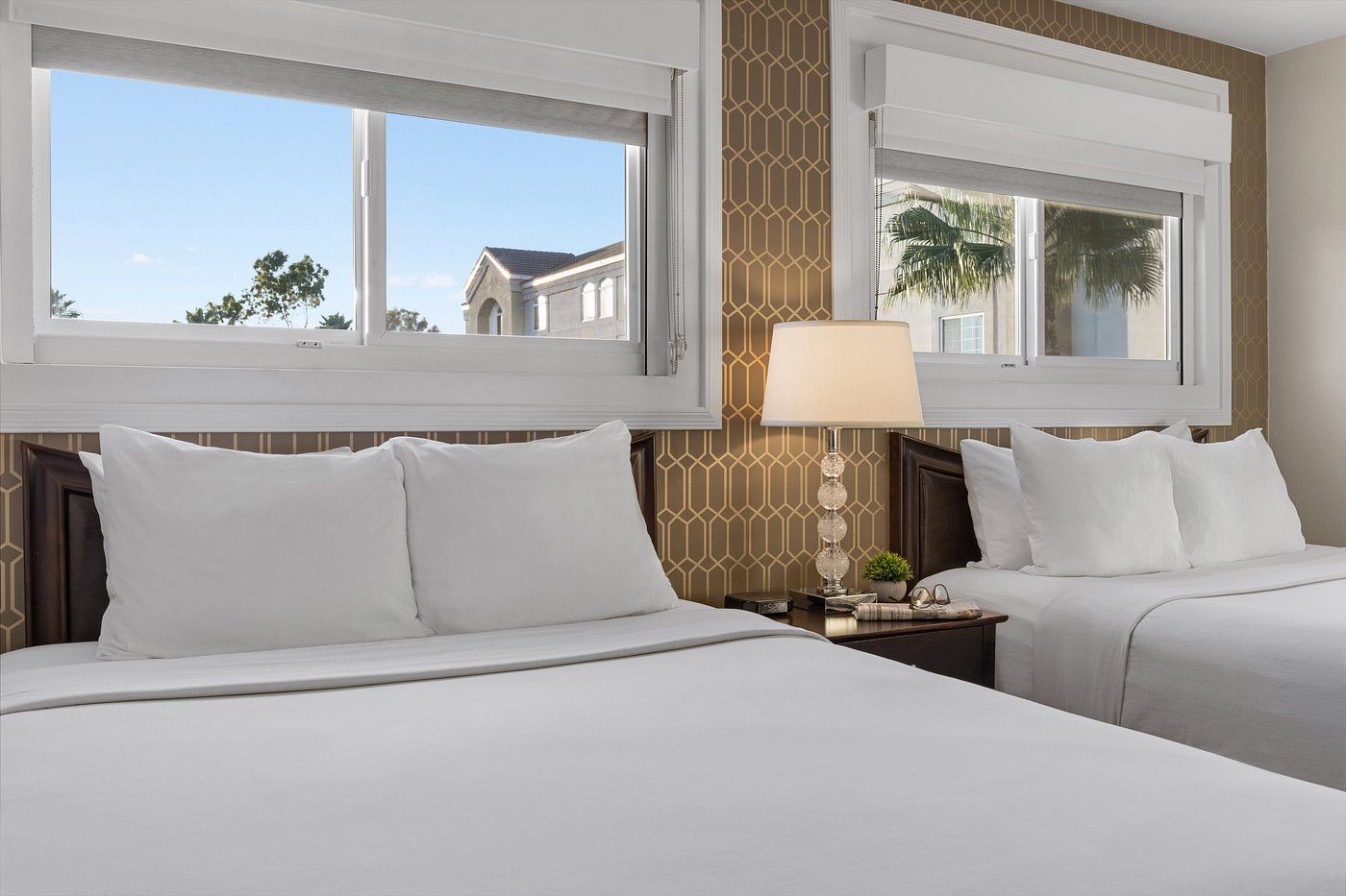 A hotel room with two beds, a nightstand with a lamp, and a view of a building through the windows.