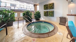 Una bañera de hidromasaje con escalera metálica, rodeada de plantas en macetas, en una habitación con suelo de baldosas y vistas a edificios.