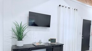 A modern living room with a flat-screen TV mounted on the wall, a dark wooden console table with decorative items, a potted plant, and a door with a white curtain. - Serenity @ Court (Sanford)