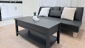 A modern living room with a dark wooden coffee table, a black sofa with white cushions, and a small potted plant. - Serenity @ Court (Sanford)