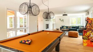 A pool table with balls, a sofa, and a colorful artwork in a bright room with large windows.