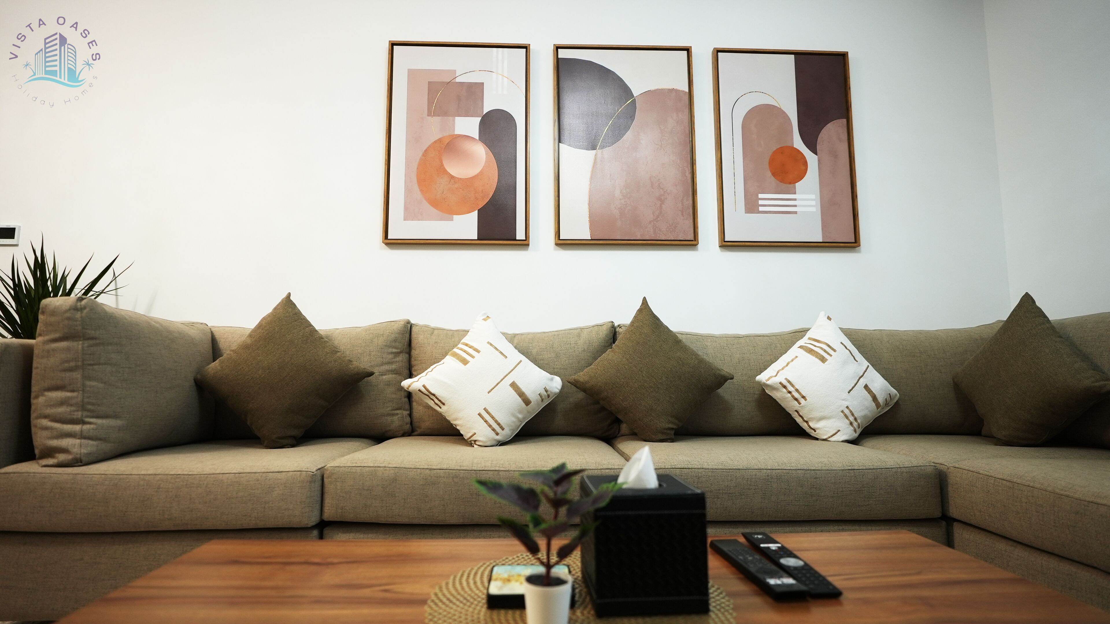 Un salon moderne avec un canapé d’angle beige, trois tableaux abstraits, une petite plante en pot et une télécommande posée sur une table basse en bois.