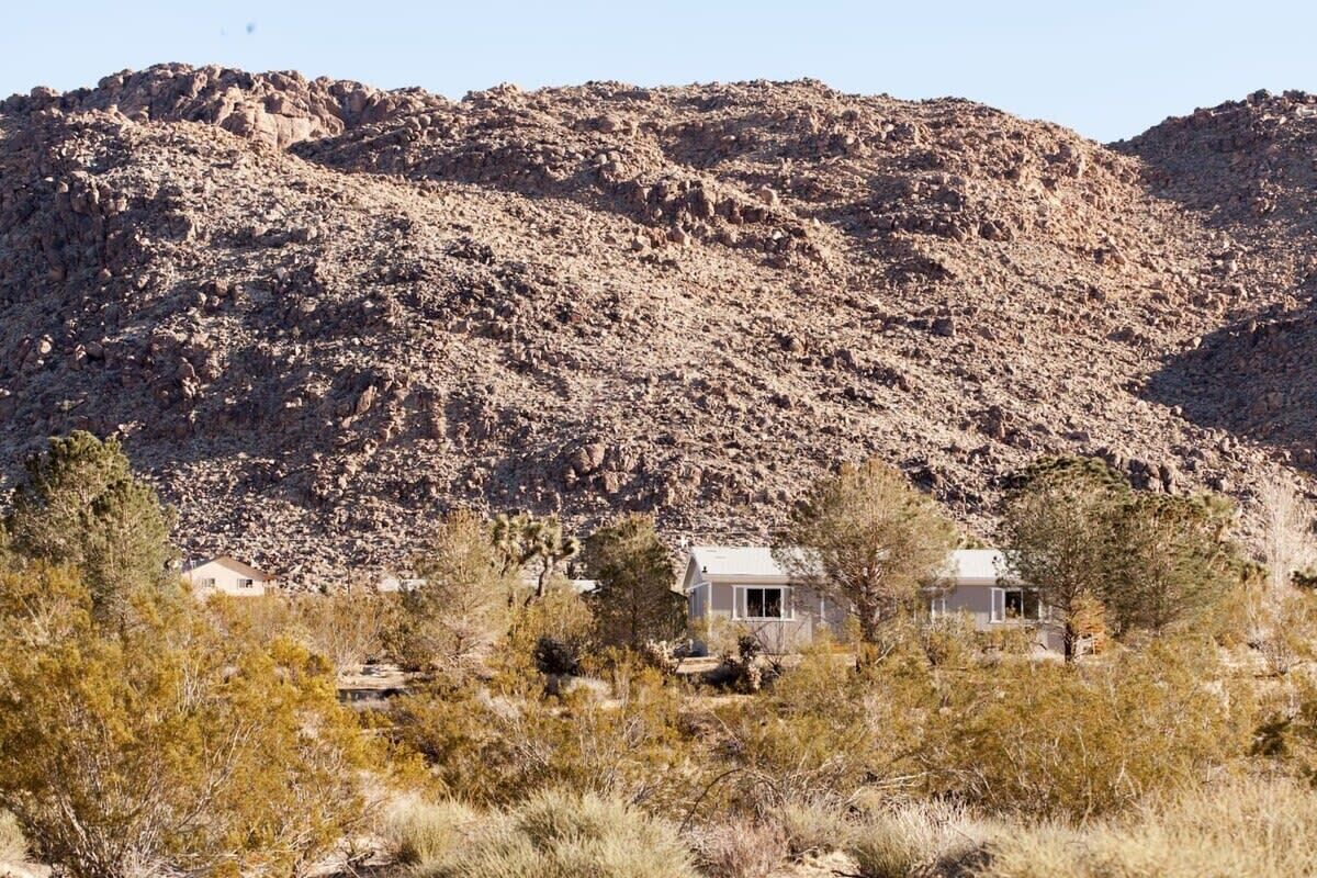 A house with a flat roof, surrounded by dry vegetation and a rocky hill in the background.