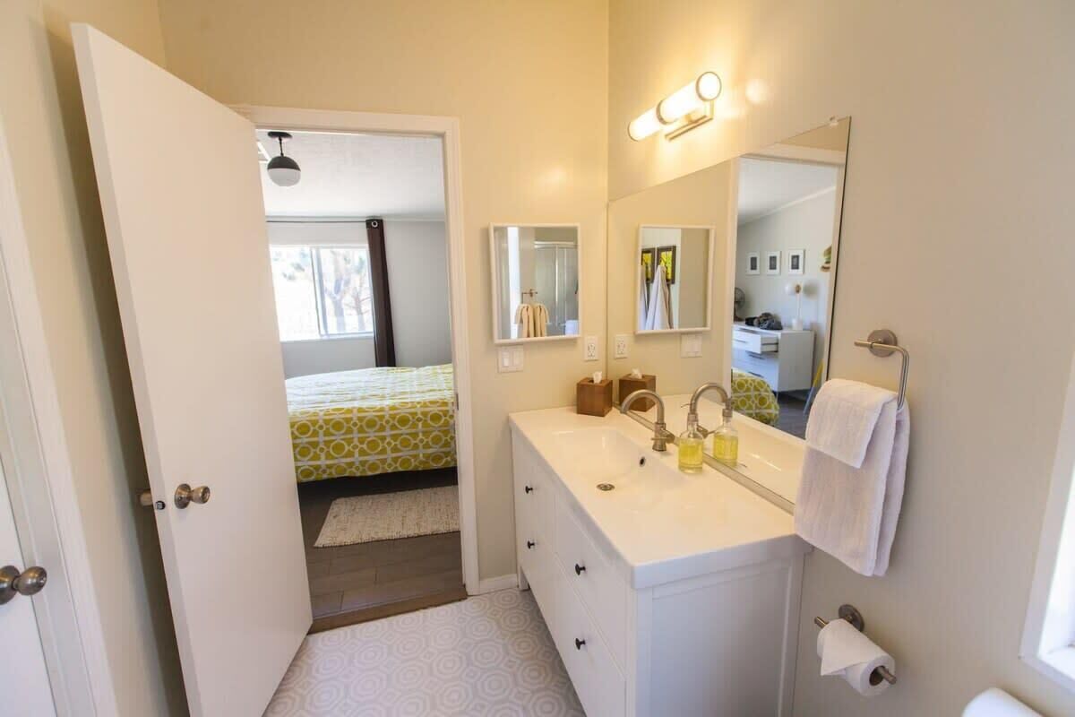 A bathroom with a white sink, mirror, and towel rack. There is a door leading to a bedroom with a bed and a window.