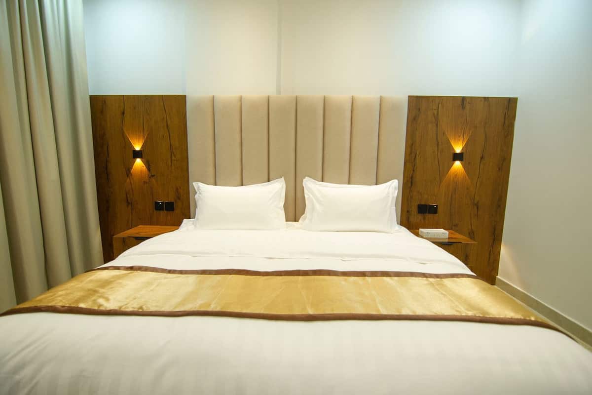 Un lit bien fait, aux couleurs beige et blanc, flanqué de têtes de lit en bois intégrant des appliques murales.