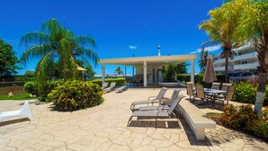 Un espacio exterior soleado con sillones de descanso, una zona de estar cubierta y palmeras.