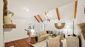Un salon spacieux avec un canapé beige, un fauteuil gris et une table basse. On y trouve un lustre, un élément décoratif rond fixé au mur et une lampe. Le salon est doté de poutres en bois au plafond et d’une grande fenêtre.