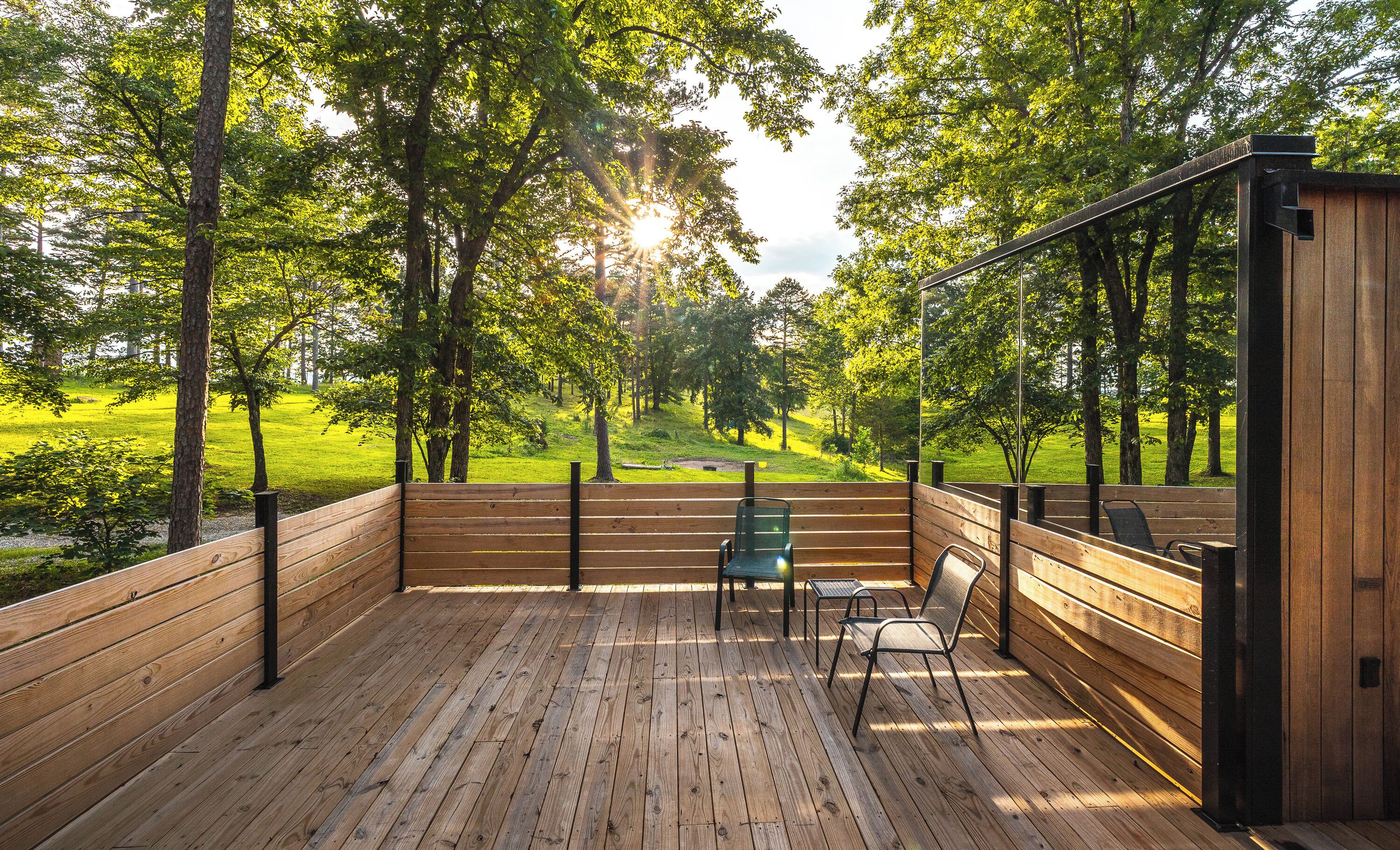 A wooden deck with benches and chairs, surrounded by trees and a view of a grassy area.