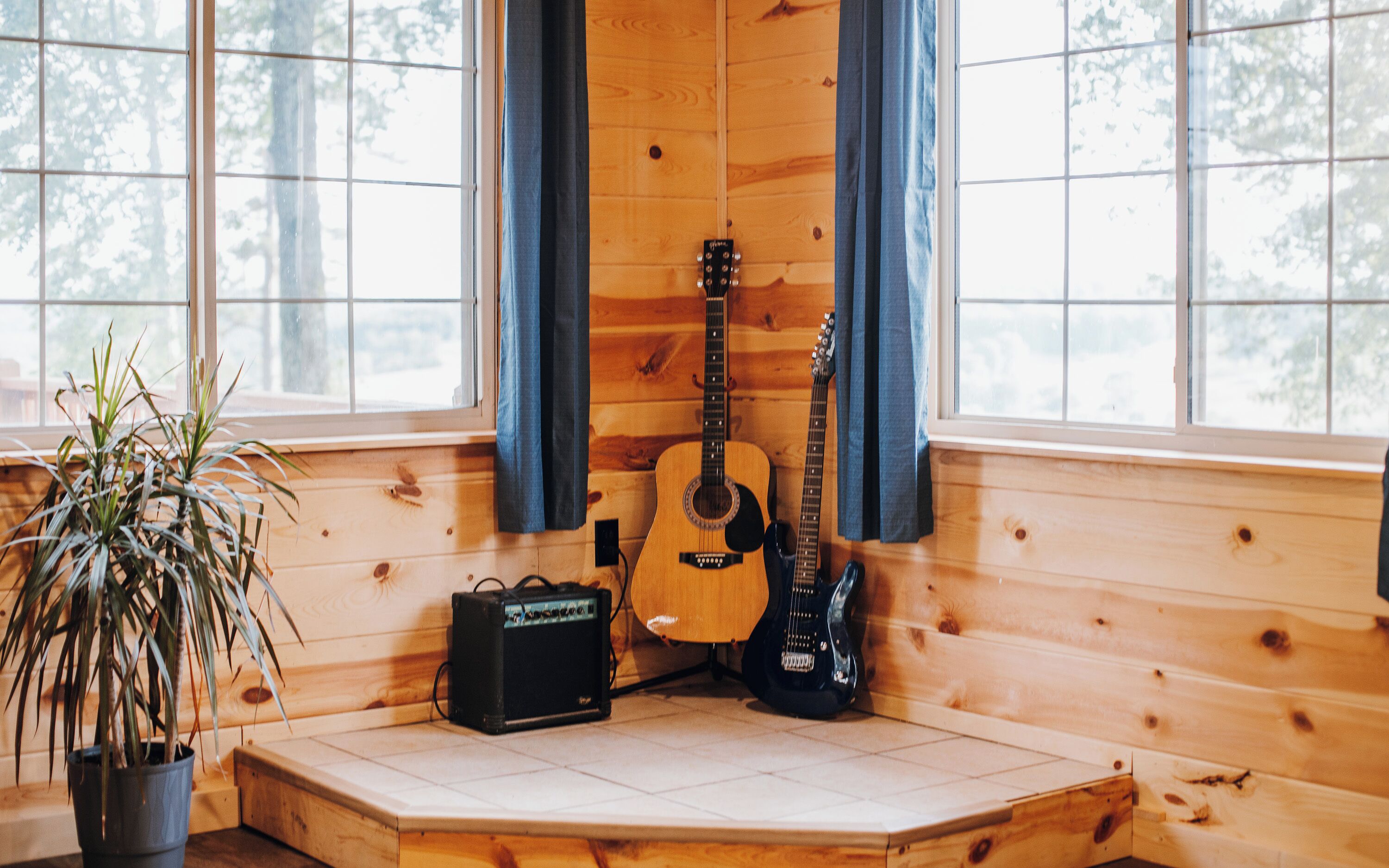 A wooden cabin interior with a guitar, amplifier, and potted plant by a window.
