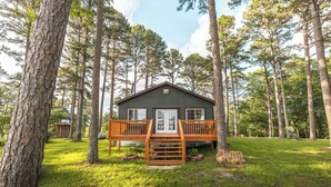 A cabin with a wooden deck and a front door, surrounded by tall trees and a grassy yard. - Floating and Glamping at Big Piney River (Licking)