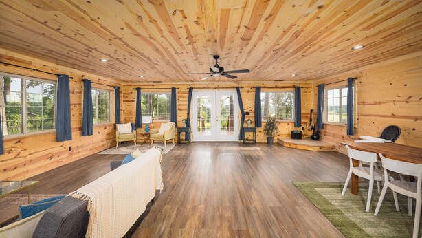 A spacious room with wooden walls and ceiling, large windows, a ceiling fan, a dining table with chairs, and a sofa. - Floating and Glamping at Big Piney River (Licking)