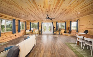 A spacious room with wooden walls and ceiling, large windows, a ceiling fan, a dining table with chairs, and a sofa. - Floating and Glamping at Big Piney River (Licking)