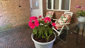 A brick exterior with a white door and windows, outdoor seating with striped cushions, and a potted plant with pink flowers. - Cozy One Bedroom Basement Apt with Free Parking (Markham)