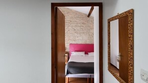 Een smalle gang met houten balken, een bed met roze beddengoed en een spiegel met houten lijst aan de muur.