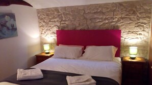 Een opgemaakt bed met witte lakens en een zwarte spreidek, met aan beide kanten nachtkastjes en lampen. De kamer heeft een achtergrond van een stenen muur en een rode hoofdbord.