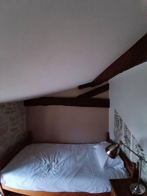 A single bed with a white mattress and a wooden headboard. There is a nightstand with a lamp on it. The room has a sloped ceiling with exposed wooden beams.