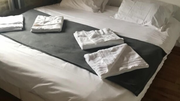 Een net opgemaakt bed met witte lakens en een donkerdeken. Er liggen opgevouwen handdoeken op het bed.