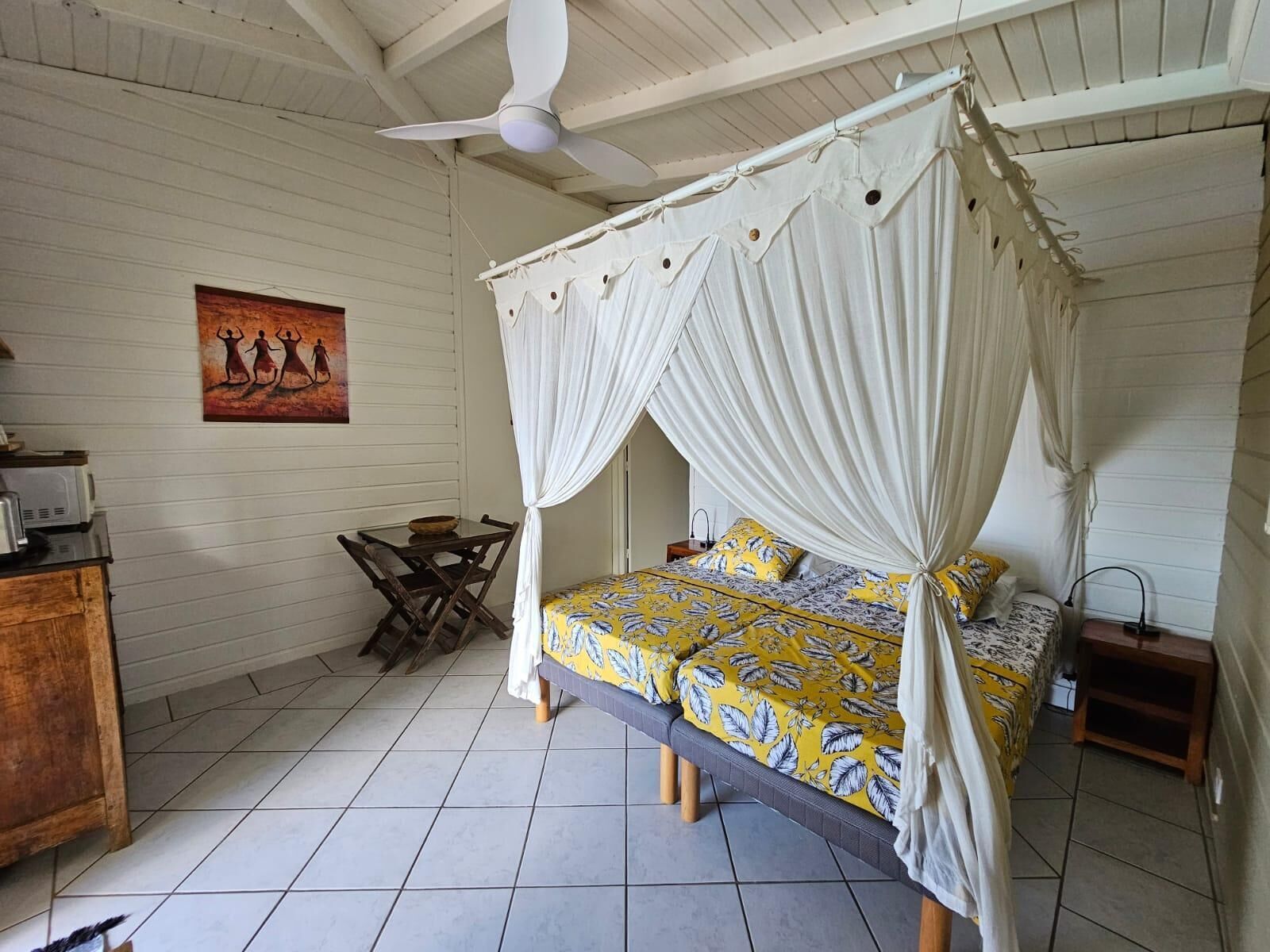 A room with a four-poster bed, a ceiling fan, a dining table with chairs, and a small wooden cabinet.