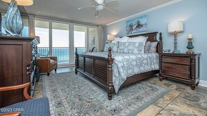 A bedroom with a large bed, a ceiling fan, a nightstand with a lamp, a dresser, and a view of the outdoors through a sliding door. - Dunes of Panama 1104e (Panama City Beach)