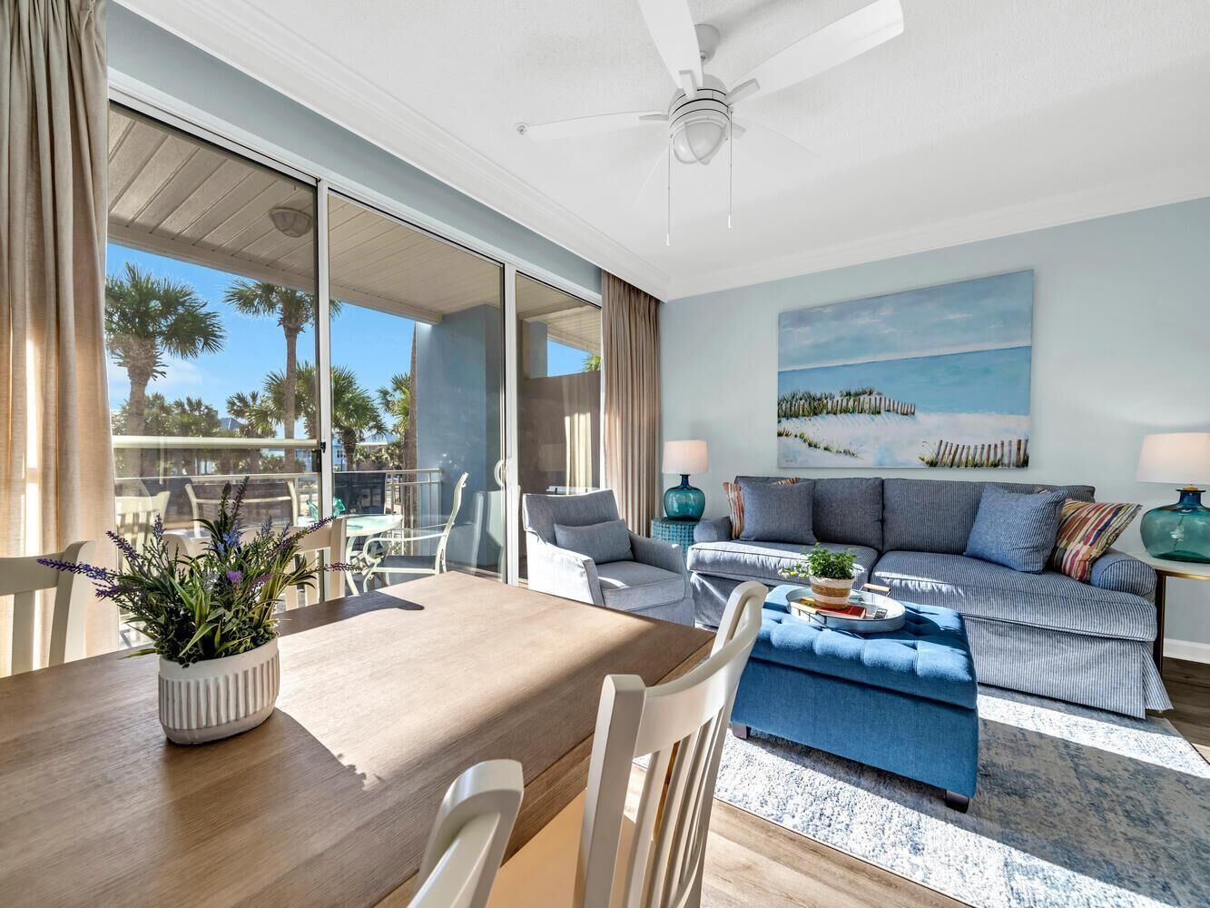 A modern living room with a sofa, a dining table with chairs, and a large window with a view of palm trees.