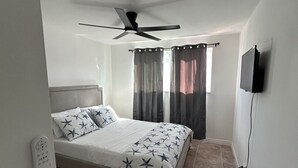 Une chambre à coucher avec un lit, un ventilateur de plafond, un téléviseur fixé au mur et une fenêtre avec des rideaux.
